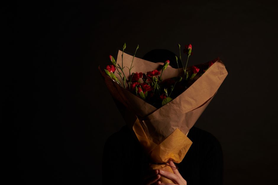 A person holding a bouquet of fresh red and pink flowers, mostly carnations, wrapped in brown kraft paper with a yellow paper accent near the base. The bouquet is held close to their face, obscuring i