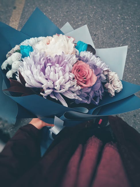 A hand holding a bouquet of fresh flowers wrapped in blue and light blue paper, featuring a large white chrysanthemum, soft pink roses, purple lisianthus, and small blue accents. The bouquet is arrang