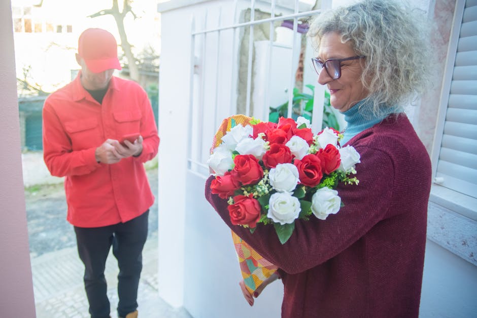 A woman with curly grey hair, wearing glasses, a blue turtleneck, and a maroon cardigan, holding a vibrant bouquet of fresh red and white roses wrapped in colorful paper. She is standing outside near 