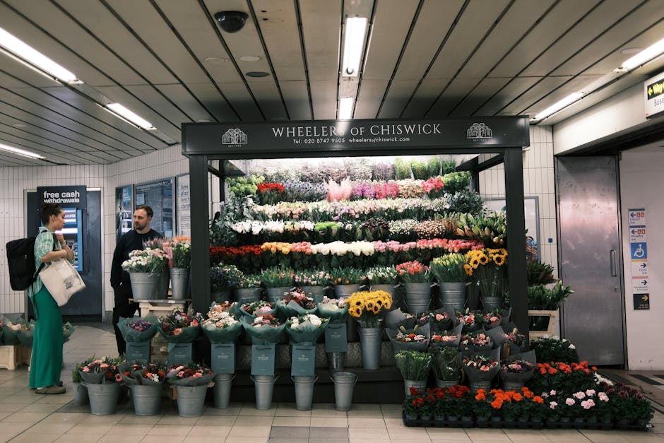A flower display stand located in an underground station environment, featuring an assortment of vibrant, freshly cut flowers arranged in metal buckets and wrapping paper. The arrangement includes pin