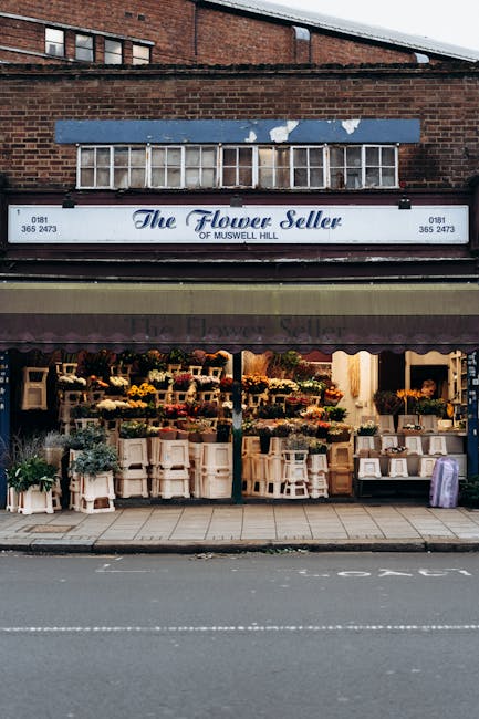 The storefront of The Flower Seller of Muswell Hill, featuring a display of fresh floral arrangements and bouquets on white shelves and stands outside the shop. The flowers include yellow, red, purple
