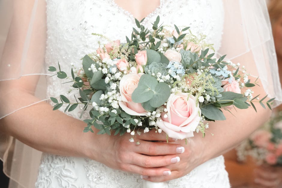 Close-up of a person in a white lace wedding dress holding a bouquet arrangement with soft pink roses, white baby's breath, green mistletoe, and dusty miller leaves, wrapped in light-colored paper wit