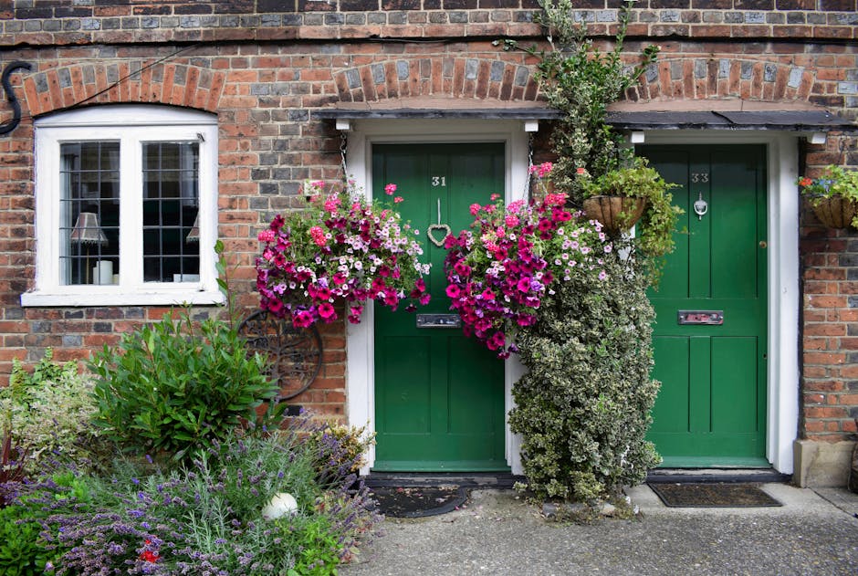 A charming brick house with two green front doors, each featuring a letterbox and door knocker, numbered 31 and 33. The area around the doors is decorated with vibrant, blooming floral arrangements, i