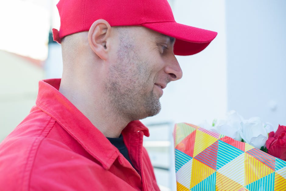 A man wearing a red cap and matching red jacket is holding a colorful floral gift bag with a geometric pattern of yellow, blue, red, and white triangles. The man has a light beard and is smiling softl