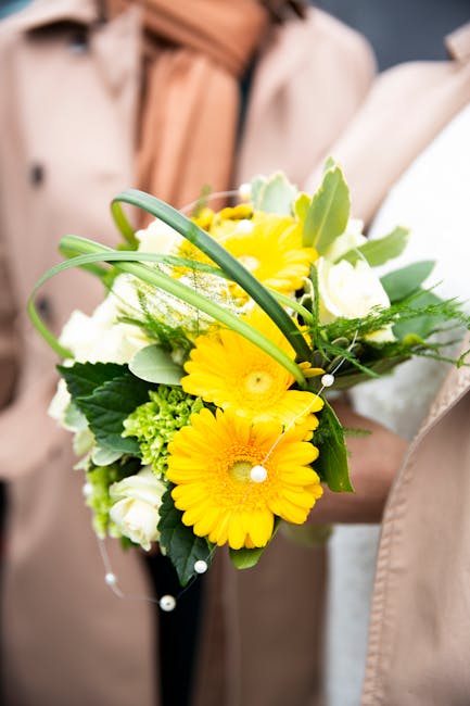 A person holding a bouquet composed of bright yellow gerbera daisies, white roses, and various green foliage, including broad leaves and smaller green accents. The bouquet is arranged in a rounded, sl