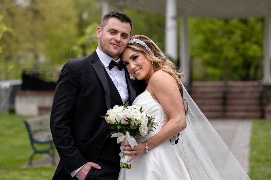 A bride and groom stand close together outdoors, smiling at the camera during their wedding ceremony. The bride wears a strapless white wedding gown with a flowing veil and holds a floral bouquet cons