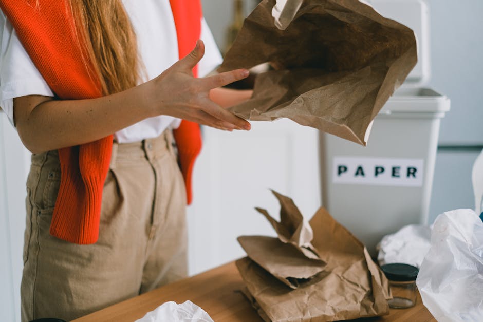 A person wearing a white shirt and an orange sleeveless sweater is holding a brown paper bag over a wooden table cluttered with crumpled brown paper and white plastic, with a white recycling bin label