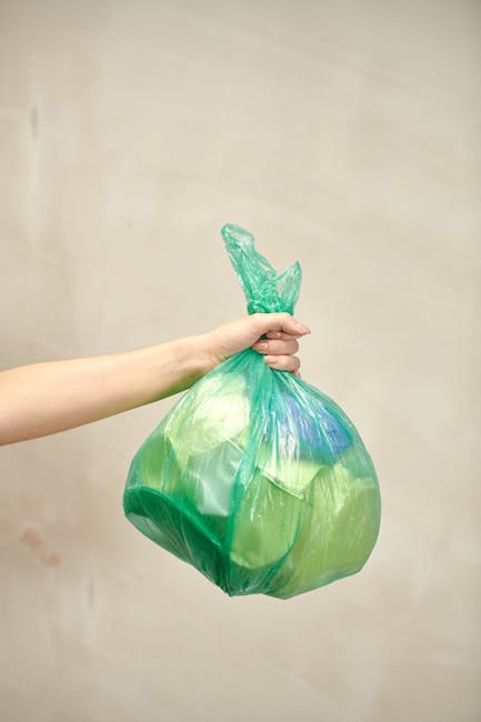 A person's hand holding a large, multicoloured plastic bag filled with waste, with the bag tied at the top and visible green and blue hues. The background is neutral and plain, emphasizing the waste d