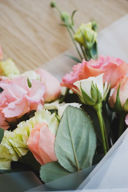 A close-up of a floral bouquet featuring soft pink and cream lisianthus flowers with ruffled petals, complemented by green leaves and stems. The flowers are arranged in a clear glass vase resting on a