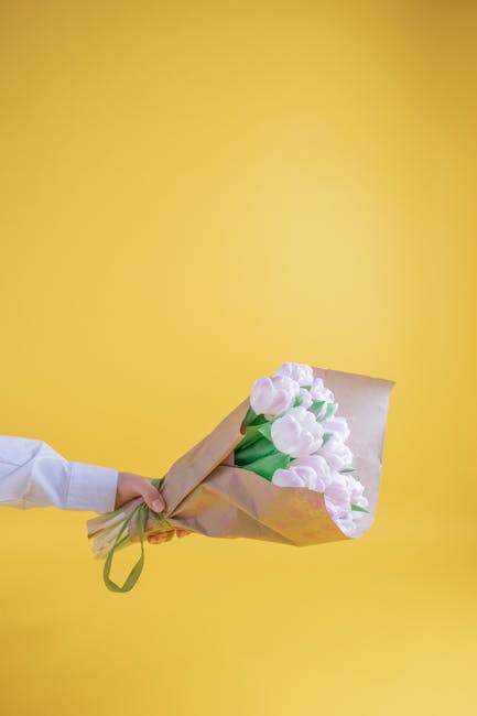 A person wearing a white sleeve holds a bouquet of light pink tulips wrapped in light brown kraft paper with a green ribbon against a solid yellow background. The tulips have smooth, rounded petals an